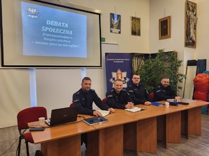 umundurowani czterej policjanci siedzący przy stole, za nimi wyświetlona prezentacja i baner KPP Kraków