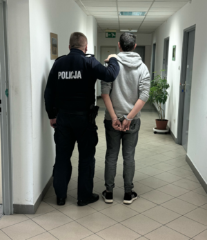 policjant stojący na korytarzu komisariatu z zatrzymanym który ma załozone kajdanki na ręce trzymane z tyłu, widoczni od tyłu (2)