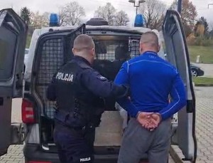 policjant wprowadzajacy do radiowozu zatrzymanego z założonymi kajdankami na ręce trzymane z tyły