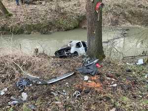 pojazd po wypadku,  leżący na brzegu rzeki, częsciowo w niej zanurzony, obok fdrzwo, obok niego leżące poodrywane części samochodu