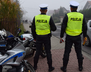 dwóch policjantów ruchu drogowego w kamizelkach odblaskowych z napisem policja stojących przy drodze, tyłem do zdjęcia, obok zaparkowane motocykle widoczne w części