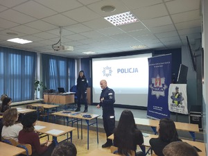 uczniowie siedzący w ławkach tyłem do zdjęcia, przed nimi dwoje policjantów, za nimi prezentacja i baner KPP w Krakowie..