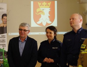 dwoje policjantów i przedstawiciel starostwa, w tle wyświetlona prezentacja z logo powiatu krakowskiego