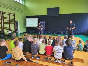 dzieci siedzące na sali gimnastycznej podczas prelekcji, przed nimi dwoje policjantów z lizakiem i kajdankami w rękach i wyświetlona prezentacja oraz baner KPP Kraków