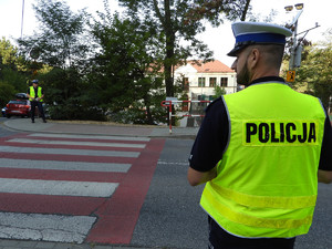 policjant stojący tyłem do zdjęcia, przodem do przejścia dla pieszych, po drugiej stronie stojący drugi policjant, w tle szkoła