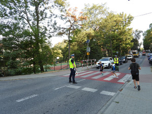 policjanci stojący przy przejściu dla pieszych, przechodzące przez przejście dzieci, a przed przejściem korek stojących pojazdów.