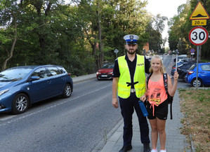 policjant stojacy na chodzniku przy drodze z dziewczynka trzymającą w ręku opaskę odblaskową