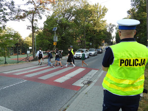 policjant stojący przy przejściu dla pieszych, kolejny policjant po przeciwnej stronie drogi w tle, przechodnie oraz stojące przed przejsciem pojazdy