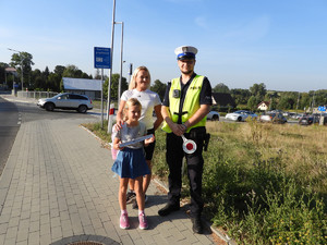 policjanmt stojacy z kobietą i dziewczynką trzymającą odblask, przy przejściu dla pieszych