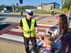 policjant wręczajacy odblaski dziewczynkom przy przejściu dla pieszych, w tle drugi policjant i szkoła