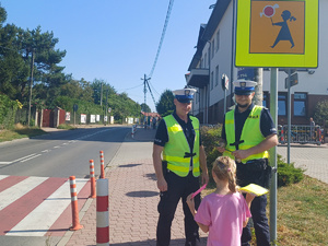 policjanci stojący przy przejściu dla pieszych, przed nimi dziewczynka stojąca tyłem do zdjęcia trzymajaca w ręku kamizelkę i opaskę odblaskową, w tle szkoła