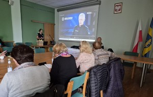 policjantka stojąca przy mikrofonie podczas prowadzenia prelekcji, obok wyświetlona prezentacja i siedzący przed nią seniorzy