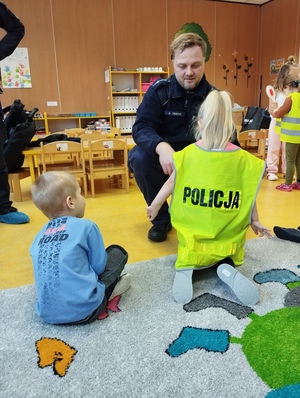 policjant zakładający kamizelkę odblaskową dzeciecku podczas prelekcji w przedszkolu, w tle inne dzieci