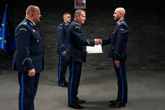 Czterech policjantów w umundurowaniu galowym. Dwóch z nich stoi naprzeciw siebie i podaje sobie dłoń.