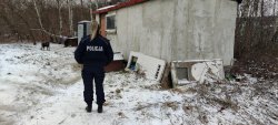 policjantka stojąca tyłem, przed nią stara zamieszkała przyczepa cempingowa, toi toi  oraz biegający pies