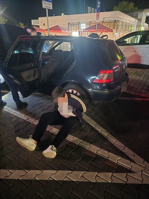 Zatrzymany mężczyzna siedzący na ziemi z kajdankami oparty o niebeski pojazd.jpg