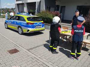 umundurowany policjant obok oznakowanego radiowozu przed nim dwojka dzieci w policyjnych ubraniach