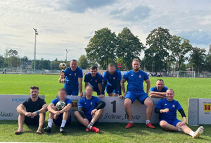 mężczyźni w niebieskich strojach sportowych na boisku piłkarskim jeden z nich trzyma puchar za I miejsce w turnieju