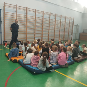 umundurowany policjant na sali gimnastycznej, przed nim siedzące siedzi na materacach podczas wykładu