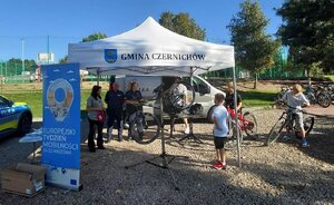 biały namiot z napisem gmina Czernichów, obok stoi radiowóz policyjny, pod namiotem stoją umundurowani policjanci z odiwedzającymi stosiko, wokół dużo rowerów