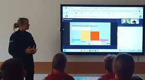 umundurowana policjantka obok dzieci siedzące w ławkach, przed nimi ekran z prezentacją