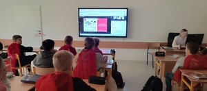 dzieci siedzące w szkolnych ławkach przed nimi ekran wraz z prezentacją