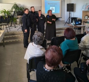 trzech umundurowanych policjantów z kobietą w czarnej sukience przed nimi siedzący seniorzy