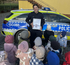 uśmiechnięta umundurowana policjanta trzyma dyplom obok oznakowany radiowóz a przy nim dzieci