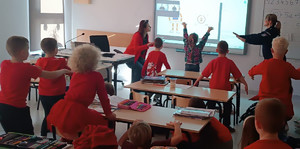 umundurowana policjantka wraz z dziewczynką patrzą na prezentację i podnoszą ręce do góry pozostałe dzieci siedzą w ławkach