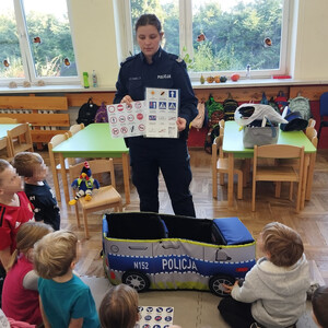 umundurowana policjantka pokazuje dzieciom znaki drogowe, dzieci siedzą na podłodze
