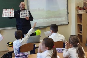 umundurowany policjant pokazuje dzieciom siedzącym w ławkach znaki drogowe