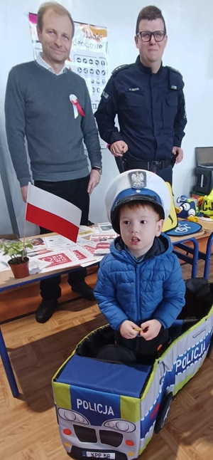 umundurowany policjant obok niego mężczyzna i dziecko w stroju policjanta i w zabawkowym radiowozie