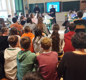 dwóch umundurowanych policjantów za nimi ekran z prezentacją, dzieci siedzą w ławkach