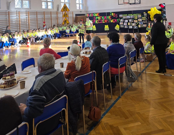 sala gimnastyczna na niej dzieci ubrane w kamizelki odblaskowe i seniorze przy stole