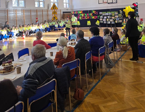 sala gimnastyczna na niej dzieci ubrane w kamizelki odblaskowe i seniorze przy stole