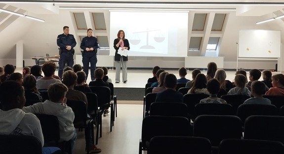 umundurowani policjanci obok kobieta z mikrofonem na sali przed nimi uczniowie na krzesłach