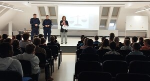 umundurowani policjanci obok kobieta z mikrofonem na sali przed nimi uczniowie na krzesłach