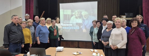 seniorzy i umundurowana policjantka na wspólnym zdjęciu obok ekran z prezentacją