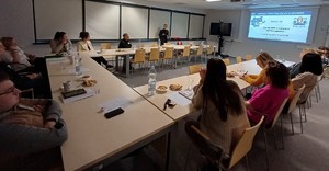 sala przy stole siedzą osoby kobieta stoi obok ekranu