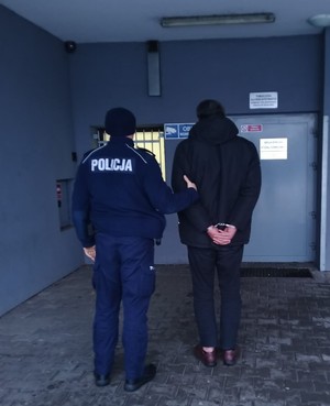 umundurowany policjant trzyma za rękę mężczyznę przed wejście do pomieszczenia dla osób zatrzymanych
