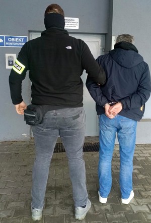 nieumundurowany policjant trzyma za rękę mężczyznę przed wejście do pomieszczenia dla osób zatrzymanych