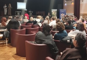 sala z fotelami na których siedzą osoby, dwie kobiety stoją z przodu z mikrofonami