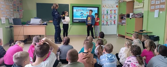 umundurowana policjantka na sali z dziećmi za nimi ekran z napisem bezpieczne zachowania