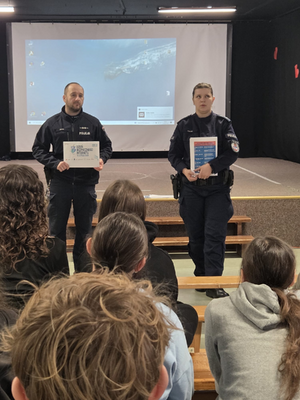 policjantka i policjant w mundurach stoją w sali przed nimi siedzą uczniowie