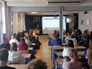 policjant w mundurze podczas prelekcji w sali przed nim siedzą uczniowie w ławkach