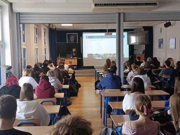 policjant w mundurze podczas prelekcji w sali przed nim siedzą uczniowie w ławkach