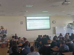 policjant w mundurze podczas prelekcji w sali przed nim siedzą uczniowie w ławkach