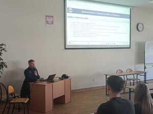 policjant w mundurze podczas prelekcji w sali przed nim siedzą uczniowie w ławkach