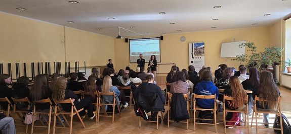 policjant w mundurze w sali podczas prelekcji przed nim siedzą uczniowie na krzesłach