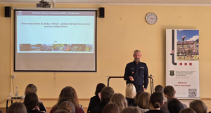 policjant w mundurze w sali podczas prelekcji przed nim siedzą uczniowie na krzesłach